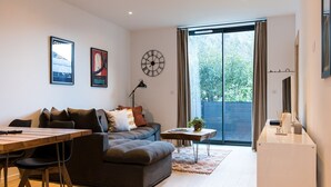 A modern living room with a sofa, a dining table, and a small side table. There is a large window with curtains, a wall clock, and two framed pictures. - Spacious Central Apartment with Terrace (Chamonix-Mont-Blanc)
