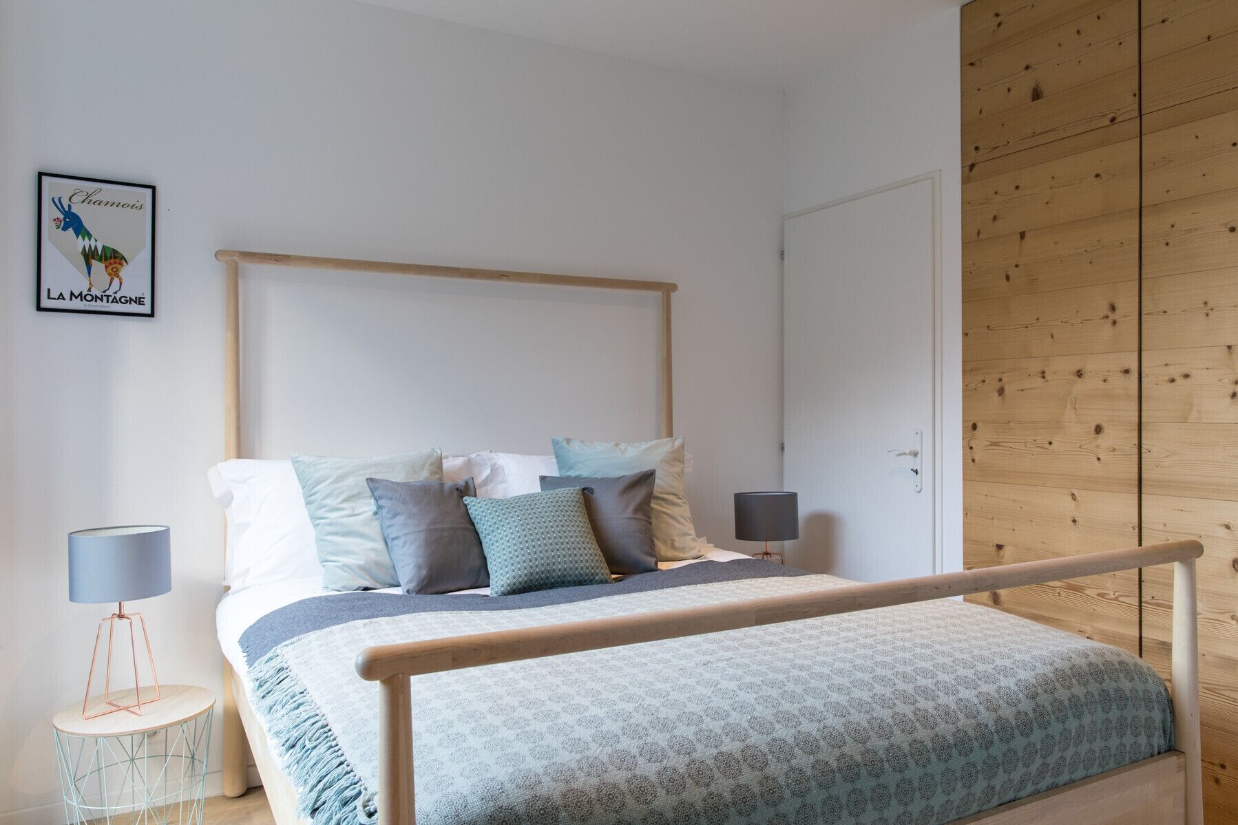 A bedroom with a wooden headboard, a bed with multiple pillows, a bedside table with a lamp, and a wall-mounted picture.