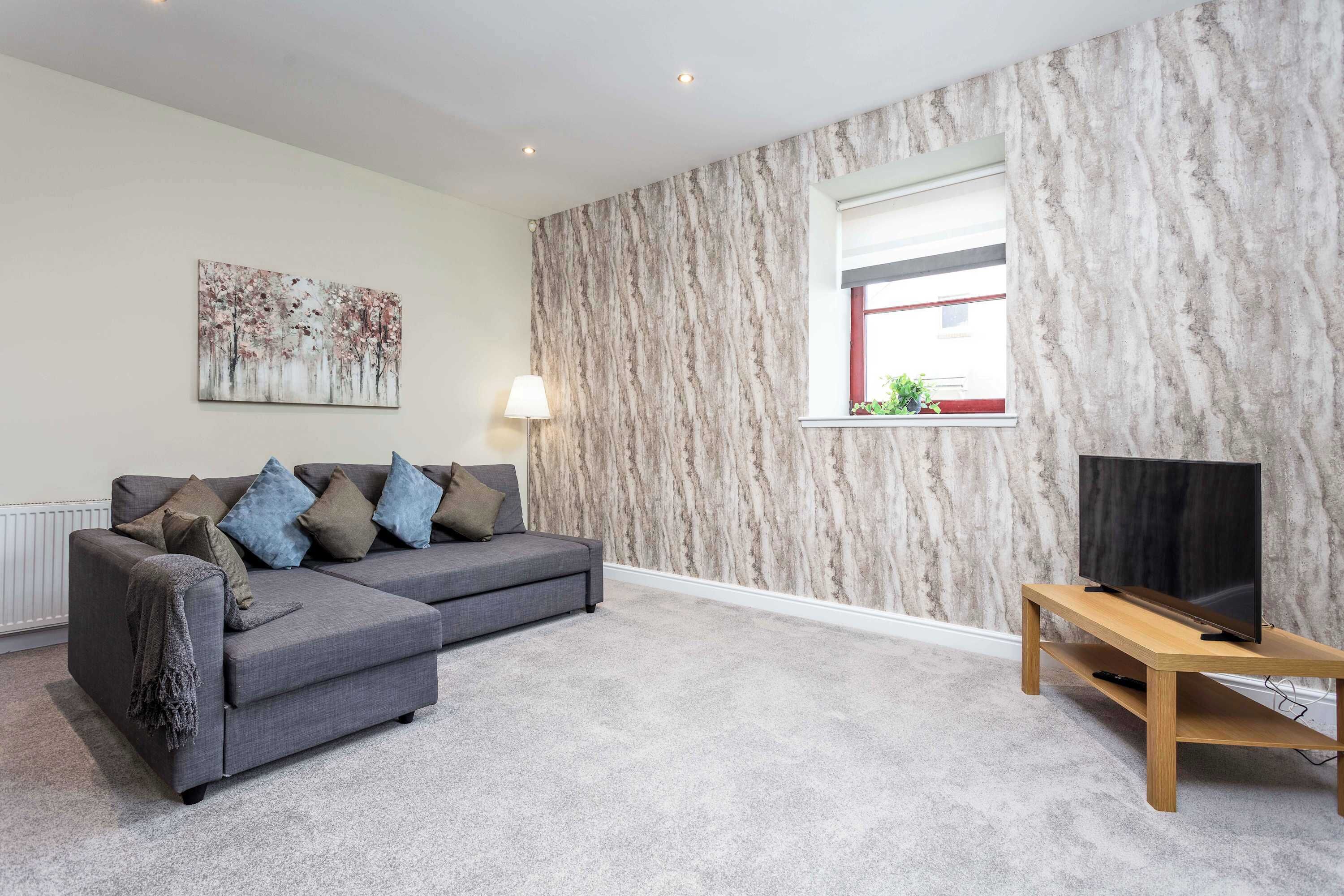 A living room with a grey sectional sofa, a flat-screen TV on a wooden stand, a marble-patterned wall, and a window with a view.