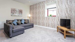 A living room with a grey sectional sofa, a flat-screen TV on a wooden stand, a marble-patterned wall, and a window with a view. - Granary Suite No3 - Donnini Apartments (Ayr)