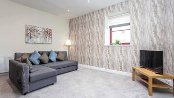 A living room with a grey sectional sofa, a flat-screen TV on a wooden stand, a marble-patterned wall, and a window with a view.