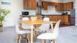 A modern kitchen with wooden cabinets, a stainless steel range hood, and a dining area with a wooden table and white chairs. - Granary Suite No3 - Donnini Apartments (Ayr)