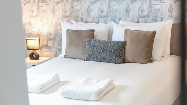 A neatly made bed with white and beige pillows, a folded white towel, and a lamp on a bedside table. - Granary Suite No3 - Donnini Apartments (Ayr)