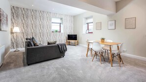 A modern living room with a grey sofa, a dining table with chairs, a TV, and a marble-patterned wall. - Granary Suite No3 - Donnini Apartments (Ayr)