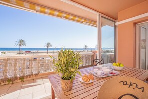 Une table en bois, sur laquelle se trouvent un panier tressé, un saladier rempli de fruits et une assiette de croissants, est placée sur un balcon donnant sur la plage.