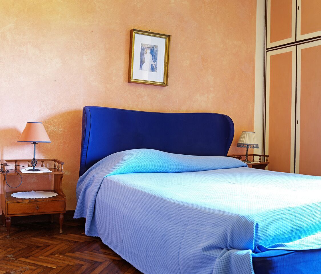 A bedroom with a blue headboard, a bed with a blue and white bedspread, a wooden nightstand with a lamp, and a framed picture on the wall.