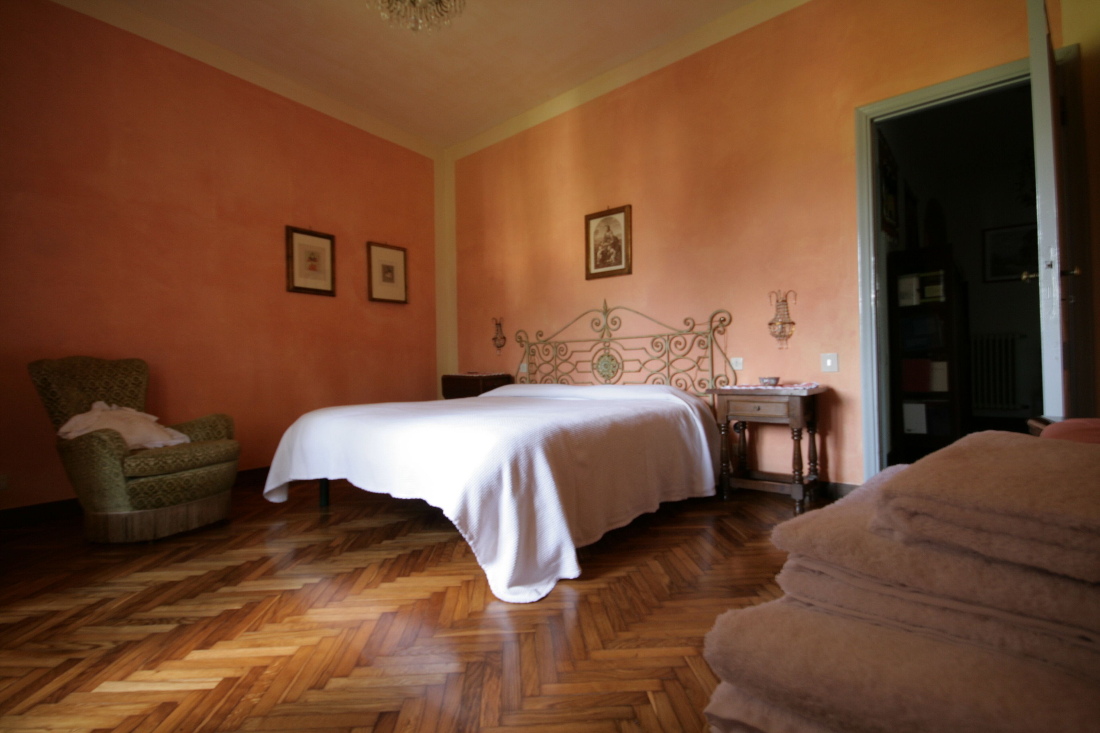 A bedroom with a wooden herringbone floor, a bed with a white bedspread, a green armchair, and a nightstand with a lamp.