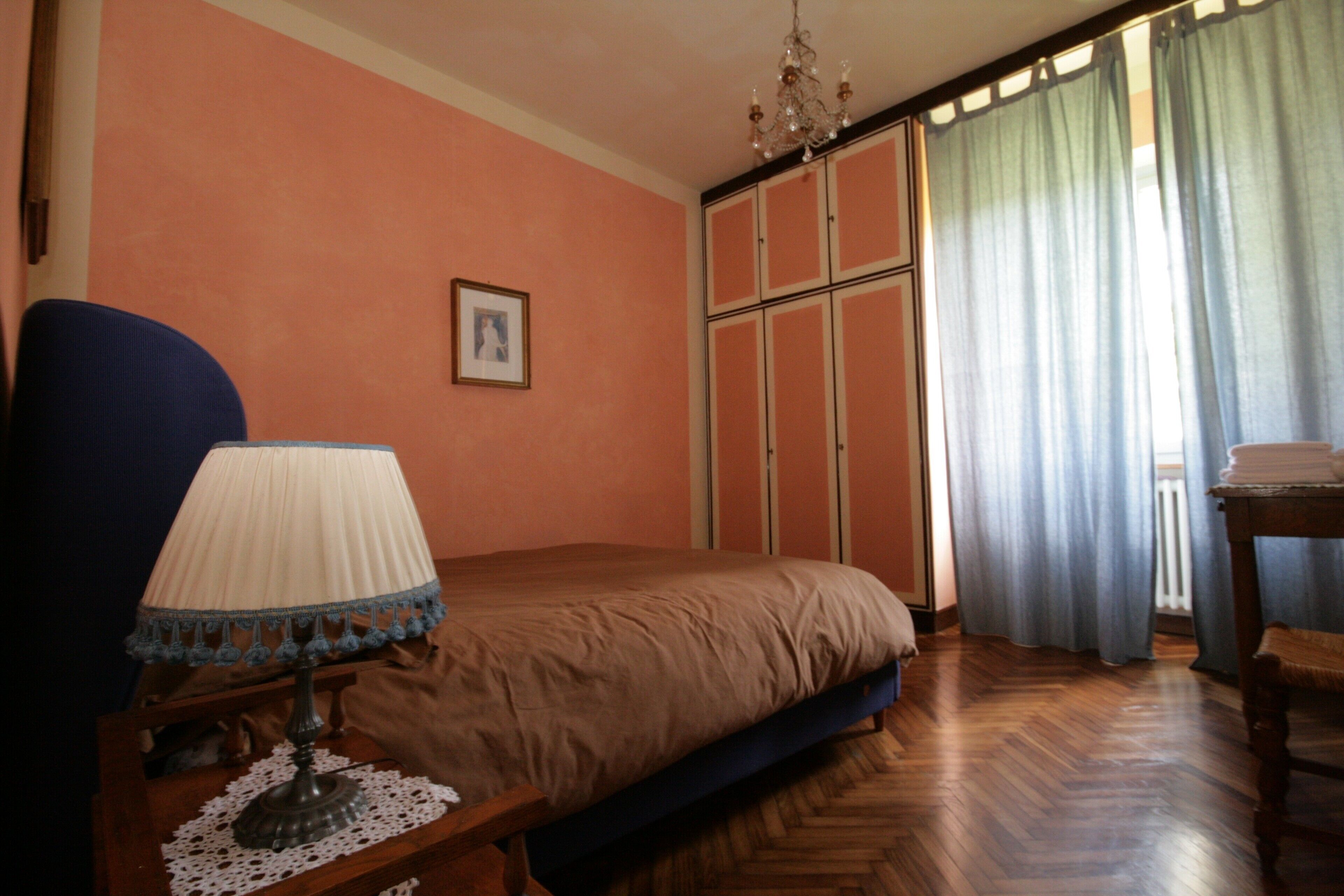 Ein Schlafzimmer mit Bett, Nachttisch mit Lampe, einem Bild an der Wand und einem Fenster mit Vorhängen.