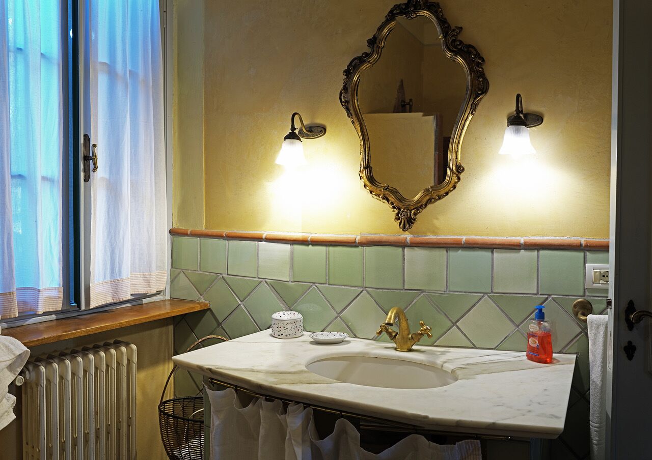 A bathroom with a white sink, gold faucet, and a large mirror with an ornate frame. There are two wall-mounted lights above the mirror and a window with sheer curtains to the left.