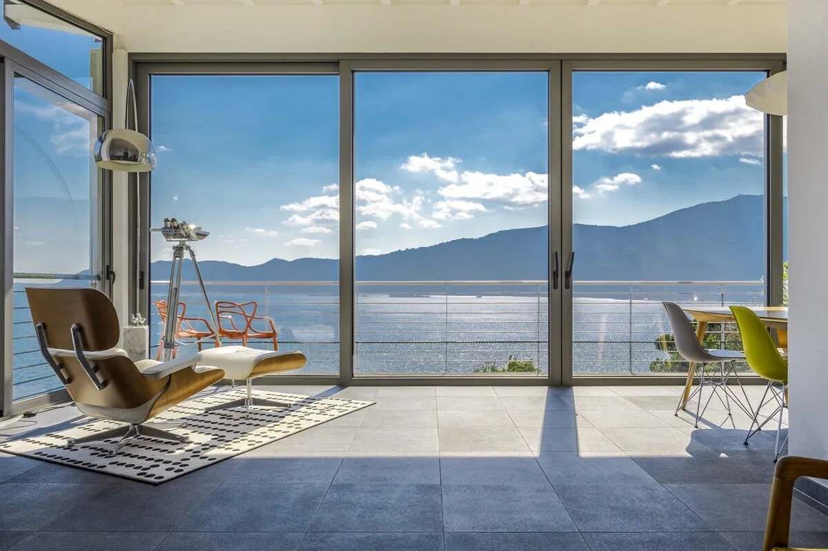 A modern room with a large sliding glass door leading to a balcony with a view of the sea and mountains.