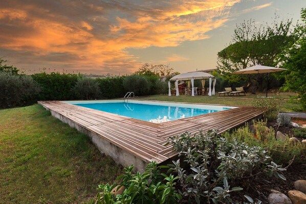 Una piscina rectangular con plataforma de madera, rodeada de césped y arbustos.