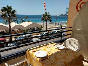 Een balkon met een tafeltje gedekt voor twee, met uitzicht op een strand met parasols en palmbomen.