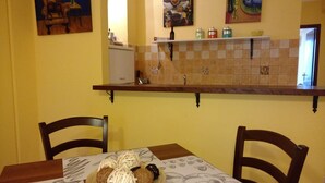 A dining area with a table set for two, chairs, a refrigerator, and kitchen shelves above the counter. - Apartment Neptune at the beach with a sea view - Letojanni, Sicily (Letojanni)