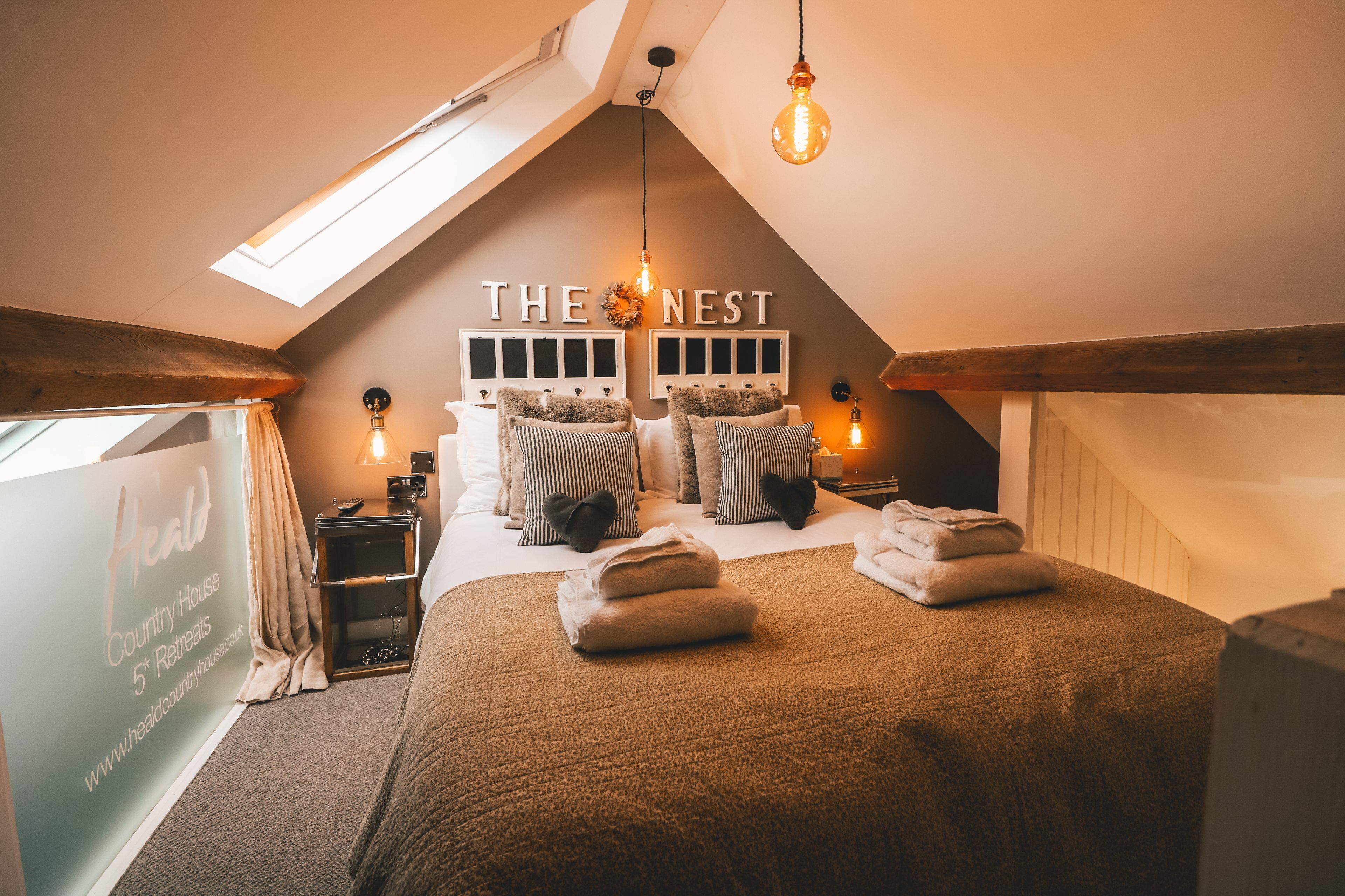 Une chambre mansardée chaleureuse et bien éclairée, avec un plafond en pente, un grand lit doté de nombreux oreillers et une pancarte sur laquelle on peut lire : « LE NID ».