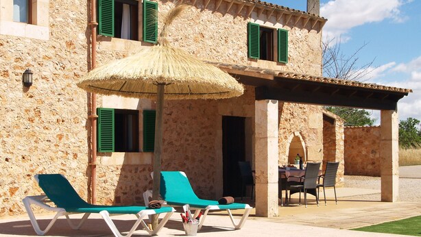 Un área junto a la piscina con sillones de descanso y una mesa, una sombrilla de paja, y un edificio de piedra con contraventanas verdes.