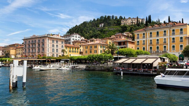 A waterfront cityscape with historic buildings, a marina, and a hill with a castle-like structure.