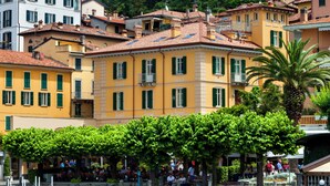 A row of colorful buildings with green shutters, a tree-lined street, and people sitting on benches.