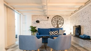 Un salon moderne avec un canapé bleu, deux fauteuils gris, une table basse noire, une décoration murale représentant un arbre et un téléviseur fixé à un mur en briques.