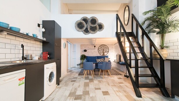 Une cuisine moderne dotée d’un lave-linge encastré, un coin repas avec un canapé bleu et une œuvre d’art murale représentant un arbre, ainsi qu’un escalier avec une rampe noire.