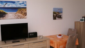 Een kamer met een televisie op een houten kast, een tafel met een oranje tafelkleed, een stoel en twee ingelijste foto's aan de muur.