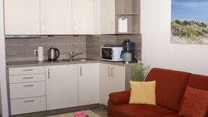 Een kleine keuken met een magnetron, koffiezetapparaat en waterkoker. Er staat een rode bank met gele kussens en een klein tafeltje met een bloevas.