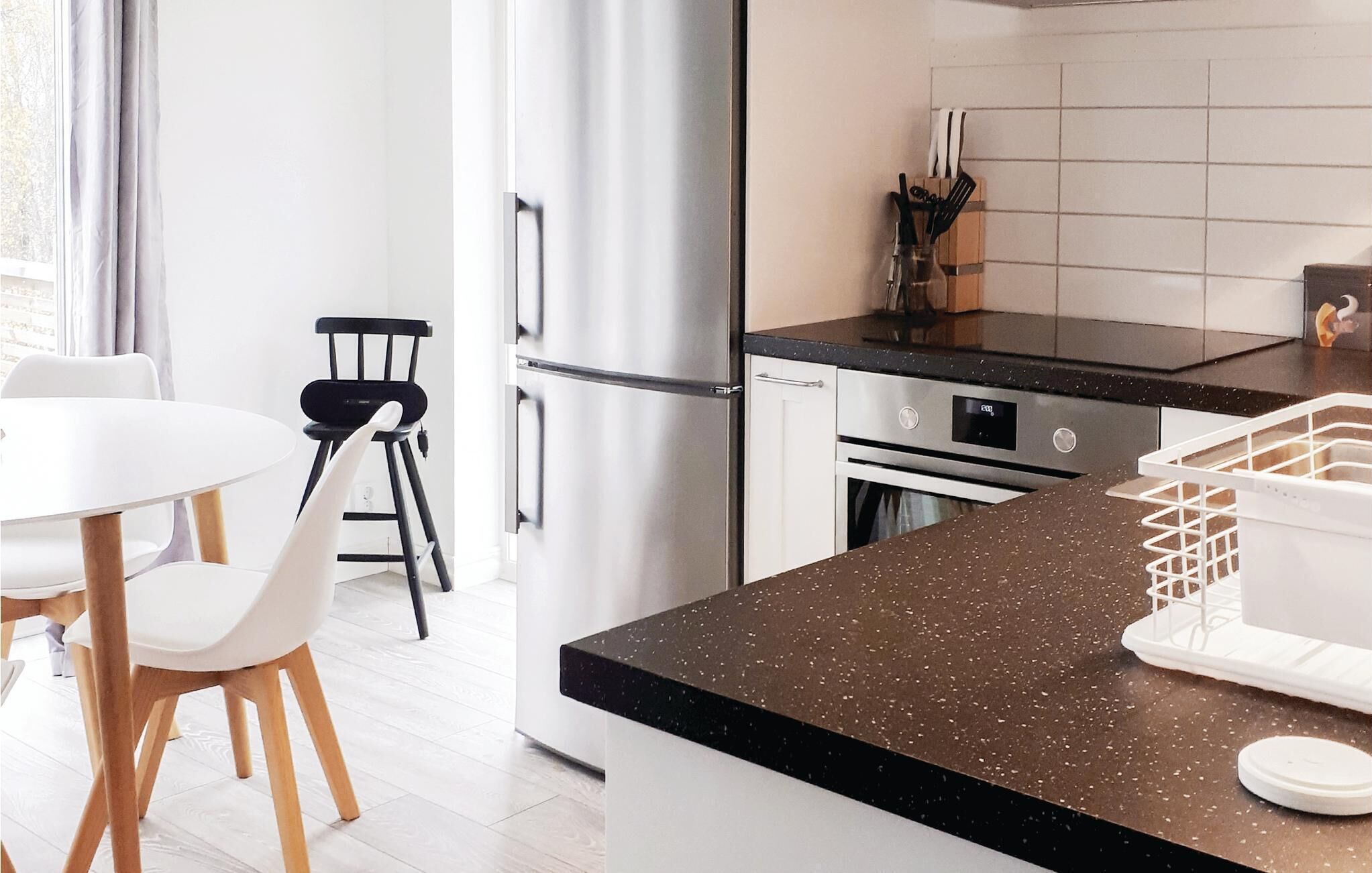 Una cocina moderna con un refrigerador de acero inoxidable, una encimera negra y un comedor con sillas blancas y una mesa.