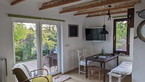 Ein Zimmer mit einer großen Glasschiebetür zu einem Garten, einem an der Wand befestigten Fernseher, einem kleinen Tisch mit Stühlen und einer weißen Bank.
