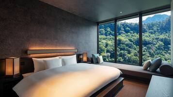 A modern bedroom with a large bed, a nightstand, a bedside table, and a window offering a view of a forested mountain range.