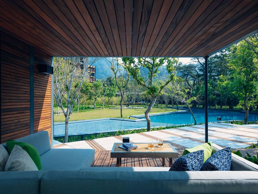 A modern outdoor seating area with a wooden ceiling, a pool, and lush greenery.