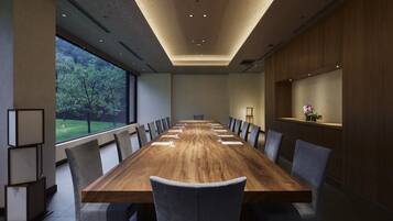 Sala de conferências com uma longa mesa de madeira, cadeiras estofadas cinzentas e vista para árvores através de grandes janelas.