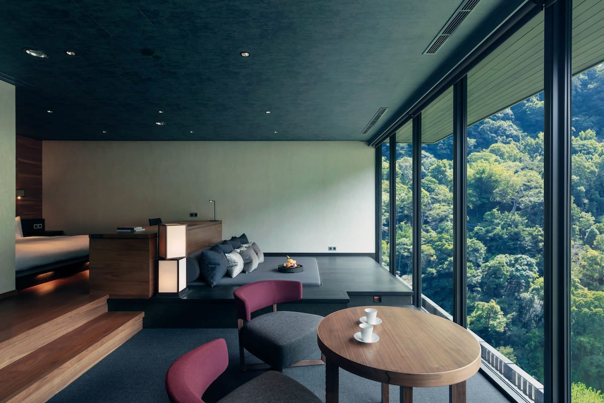 Une chambre moderne dotée d’une grande fenêtre, d’un lit, d’un bureau et d’un coin salon offrant une vue sur la forêt.