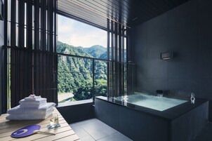 A modern bathroom with a large bathtub, a view of lush green mountains, and a stack of white towels on a wooden table.