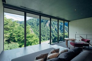 A modern room with a large window offering a view of a forested mountain. There is a sofa with cushions and a small table with chairs.
