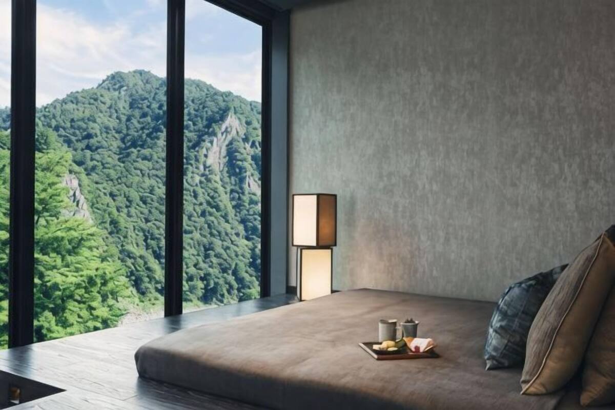 Une chambre avec une grande fenêtre, une table de chevet avec une lampe et un lit sur lequel se trouve un plateau avec divers objets.
