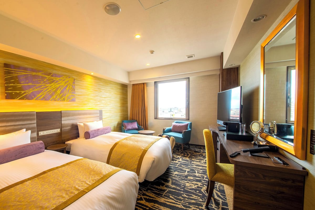 A hotel room with two beds, a desk, a chair, a TV, and a large window.