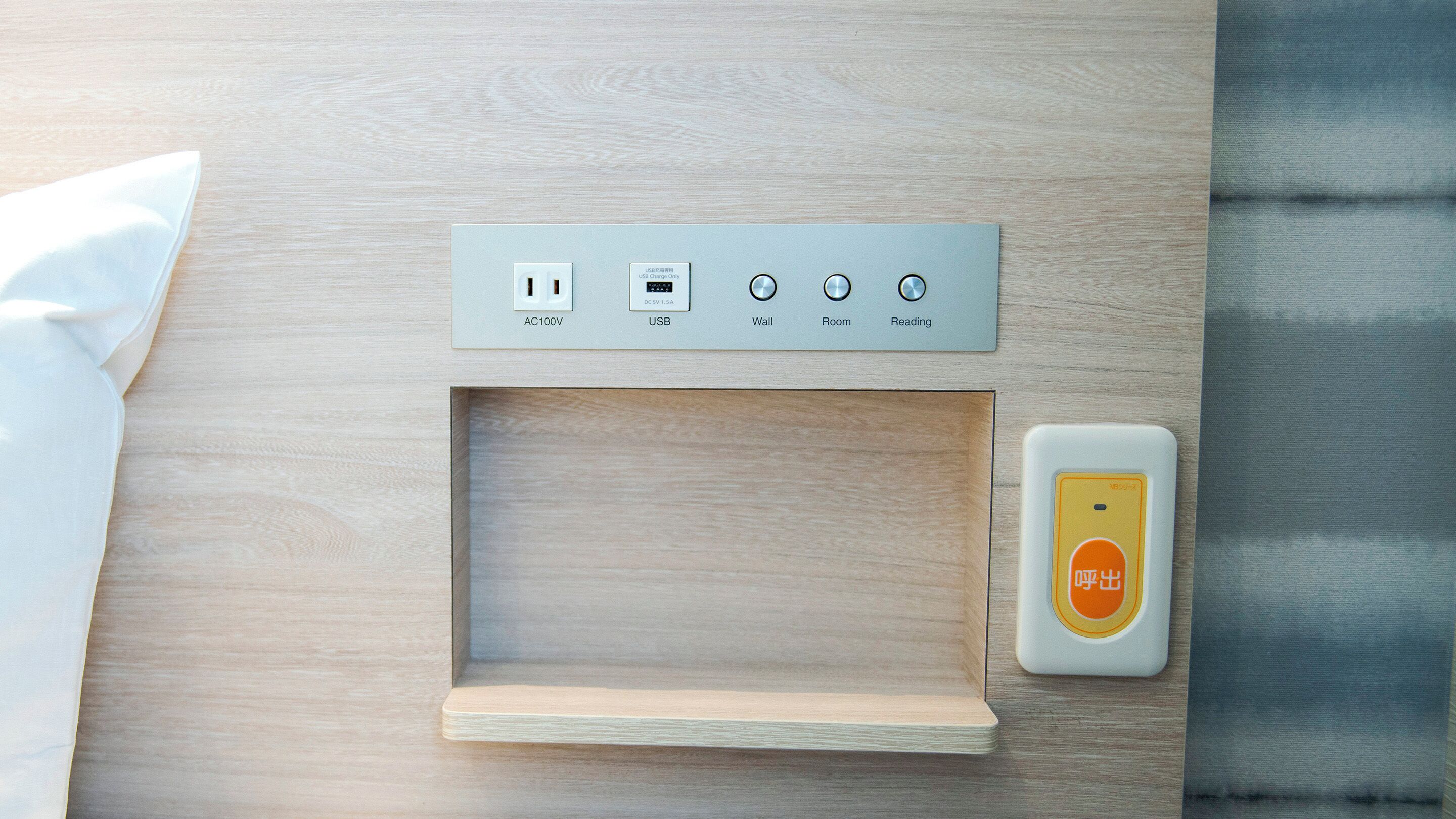 A modern, minimalist room with a wooden floor, a small wooden shelf, a wall-mounted USB charging station, and a remote control.
