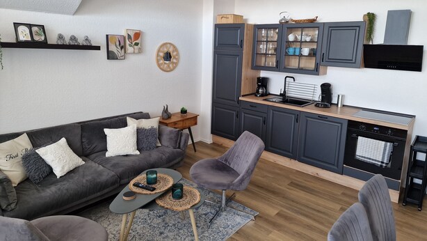 A modern living room with a grey sofa, a coffee table, and a grey armchair. A kitchen area with grey cabinets and a black stove is visible. - Holiday apartment directly on the beach of the North Sea and still central in Bensersiel (Bensersiel)