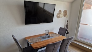 A dining area with a wooden table, four grey upholstered chairs, a flat-screen TV mounted on the wall, decorative wall art, and a window with a view. - Holiday apartment directly on the beach of the North Sea and still central in Bensersiel (Bensersiel)