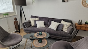 A modern living room with a grey sofa, two armchairs, a coffee table, and a floor lamp. There is a small Buddha statue, a shelf with decorative items, and a round mirror on the wall. - Holiday apartment directly on the beach of the North Sea and still central in Bensersiel (Bensersiel)