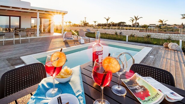 A poolside setting with a bottle of C&M wine, two glasses of red wine, a bowl of chips, and a menu on a wooden table. - Villa Ulivo for 8 People in Sicily, Pool, Sea View, Garden, Wifi and A/C (Reitani)