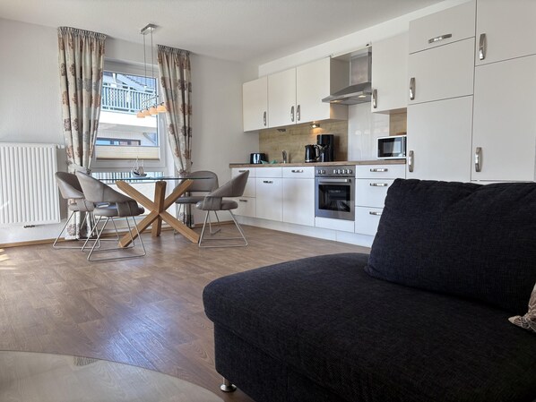 A modern kitchen with white cabinets, a built-in oven, and a microwave. There is a dining area with a glass table and two chairs. A dark sofa is in the foreground. - The "Seemöwe" in Haus Meeresglück, close to the beach and central! (Dahme)