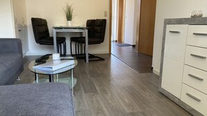 A modern living room with a grey sofa, a glass coffee table, and a small table with a plant. There is a kitchen area with a white cabinet and a wooden floor. - The "Seepferdchen" in Haus Meeresglück, close to the beach and central! (Dahme)
