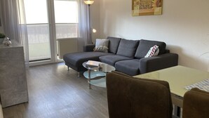 A modern living room with a grey sofa, a glass coffee table, and a dining area with a white table and brown chairs. - The "Seepferdchen" in Haus Meeresglück, close to the beach and central! (Dahme)