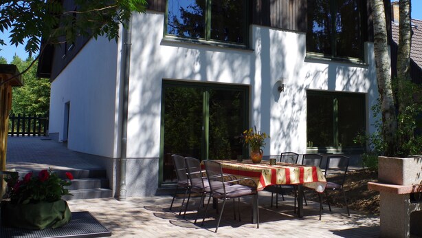 A modern two-story house with a patio area featuring a table and chairs, surrounded by trees.