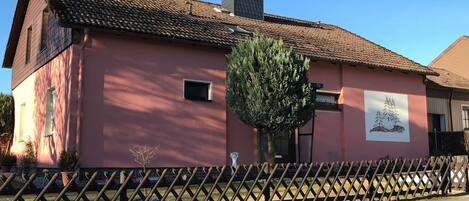 A pink building with a tiled roof, a small window, and a tree in front.