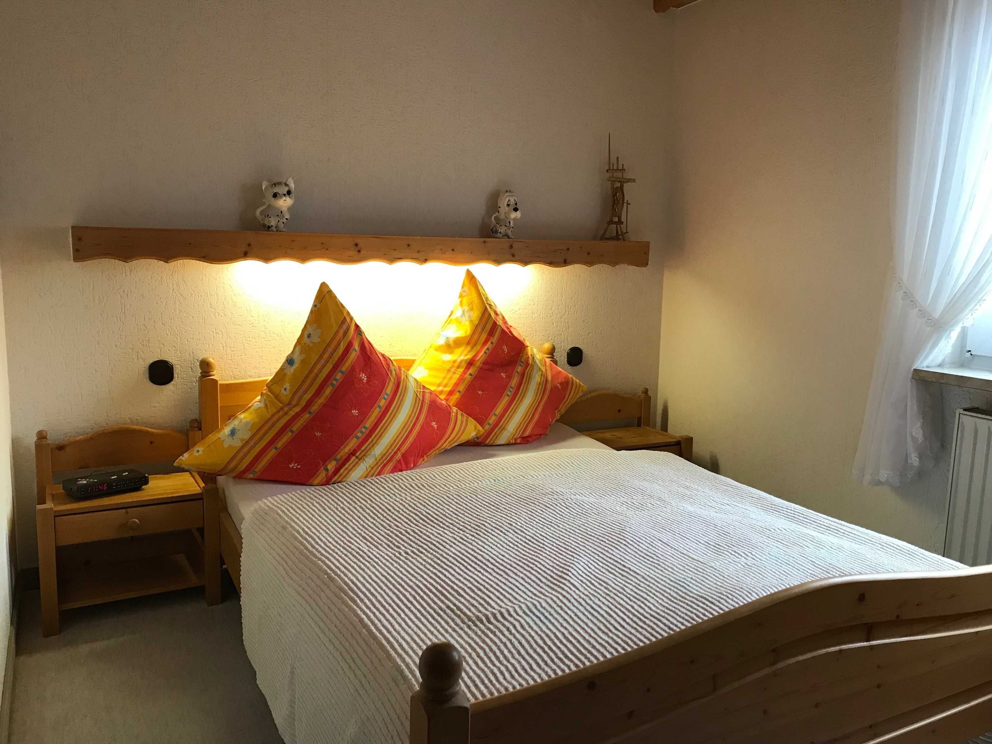 Ein Schlafzimmer mit einem hölzernen Bett, zwei Nachttischen, einem Beistelltisch, einer Wanduhr, einem Regal mit Dekoration und einem Fenster mit Vorhängen.