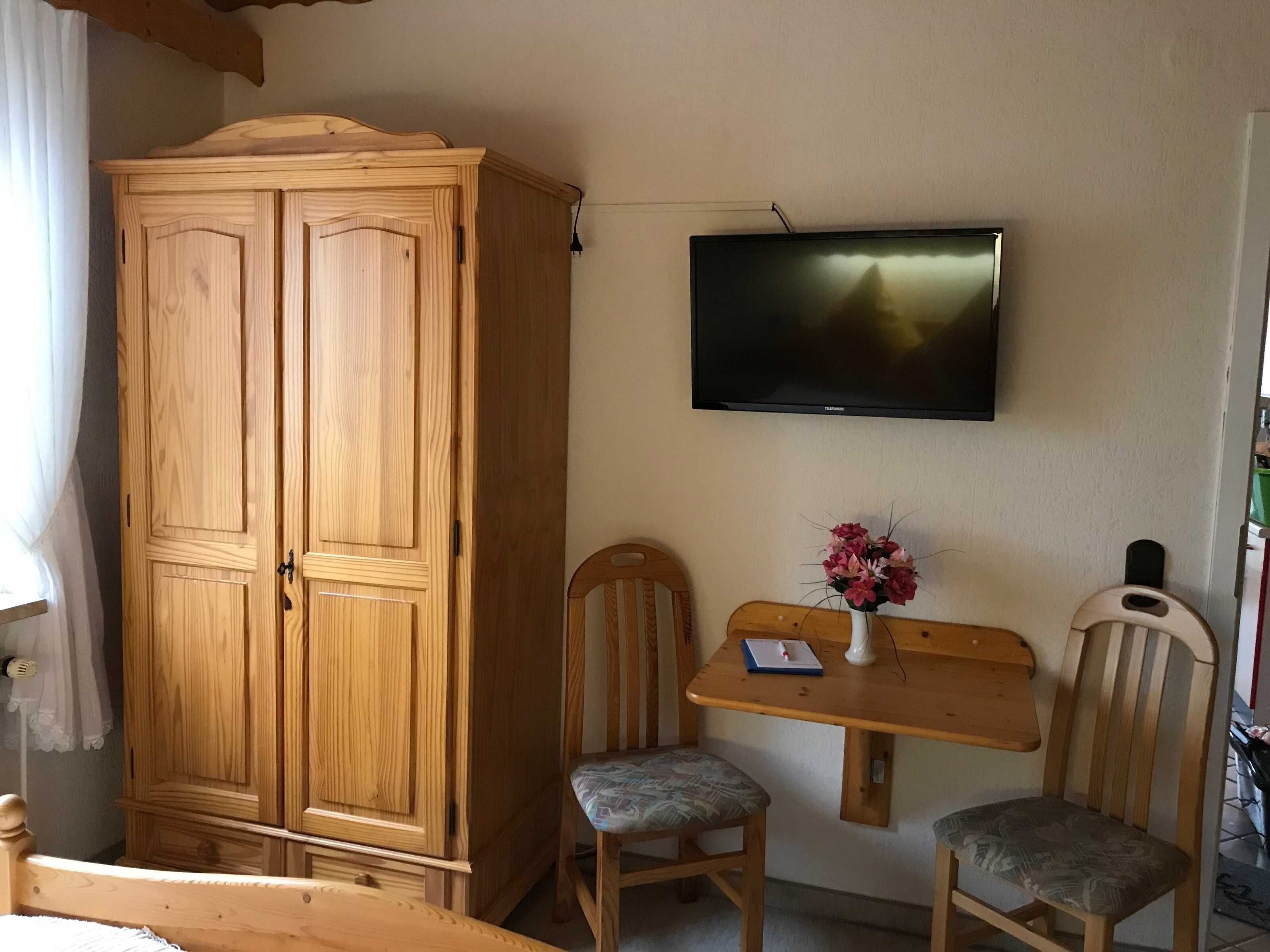 Une pièce comprenant une armoire en bois, un téléviseur fixé au mur, une petite table avec deux chaises et un vase avec des fleurs.
