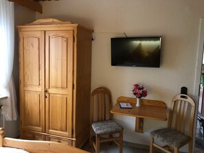 Ein Zimmer mit einem Holzschrank, einem an der Wand montierten Fernseher, einem kleinen Tisch mit zwei Stühlen und einer Vase mit Blumen.