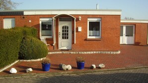 Un bâtiment en briques avec une porte blanche, deux fenêtres et un petit porche agrémenté de plantes en pot et de rochers.