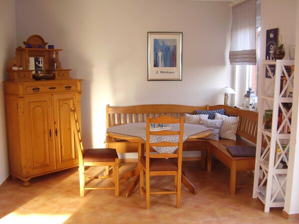 A cozy dining area with a wooden table, chairs, and a bench. A framed picture hangs on the wall, and a bookshelf is to the right.